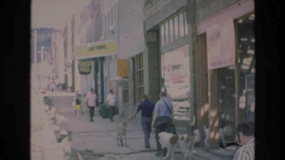 1973 Colorado Springs sidewalk view showing pedestrians near storefronts, a hanging sign, window displays, and bright downtown street activity beyond.