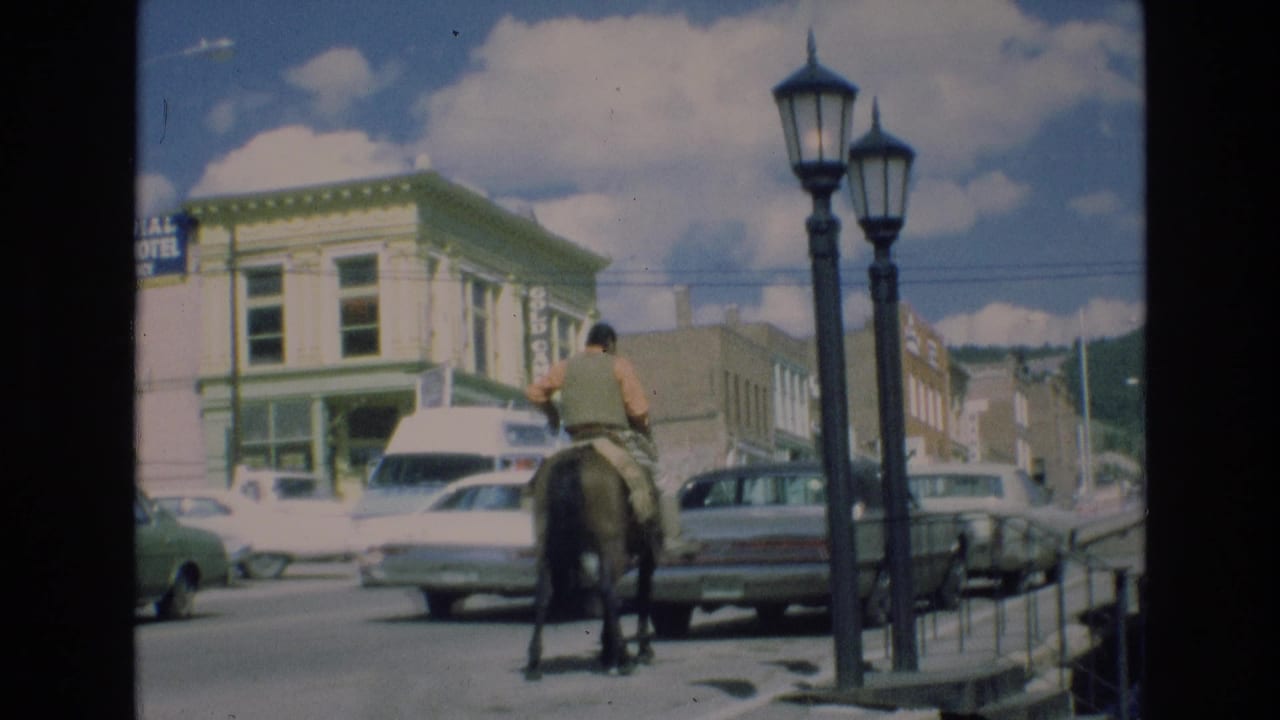 In the stated 1973 Colorado Springs footage, a man rides a horse down a commercial street with classic cars, a white van, storefronts, and tall streetlamps nearby.