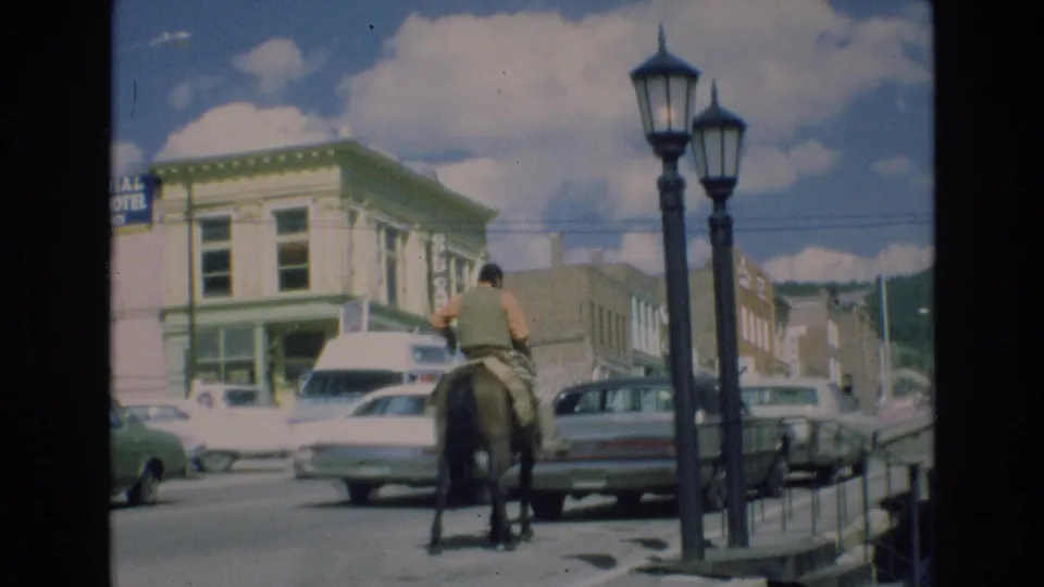 In the stated 1973 Colorado Springs footage, a man rides a horse down a commercial street with classic cars, a white van, storefronts, and tall streetlamps nearby.