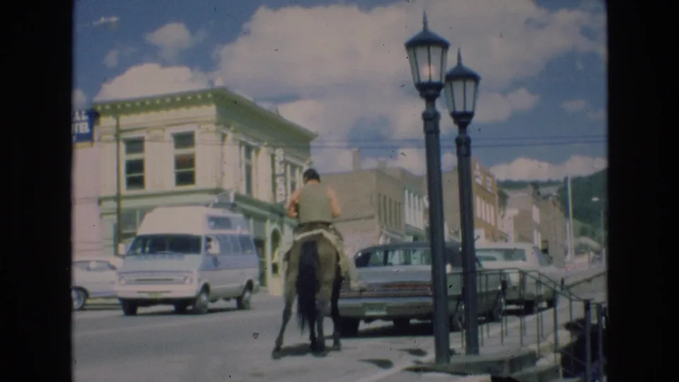 The home-movie frame shows the horse and rider farther along the Colorado Springs street, with a white van at left, older cars at right, and storefronts rising behind them.