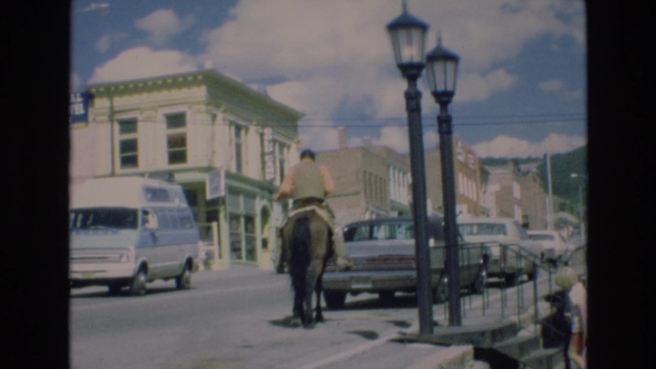 In Colorado Springs, Colorado in 1973, a man on horseback moves along a downtown street lined with parked cars, storefronts, and twin street lamps.