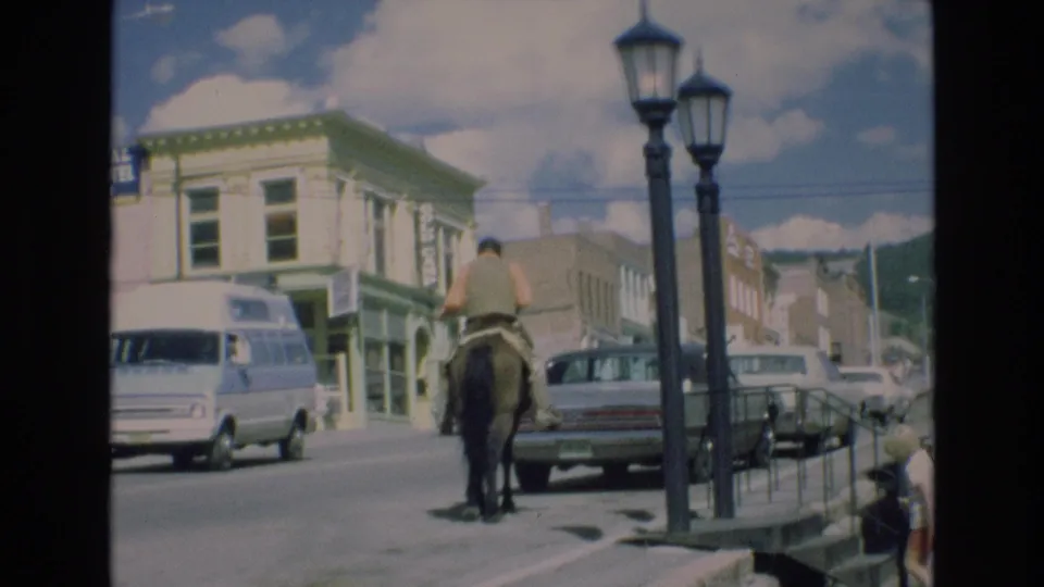 In Colorado Springs, Colorado in 1973, a man on horseback moves along a downtown street lined with parked cars, storefronts, and twin street lamps.