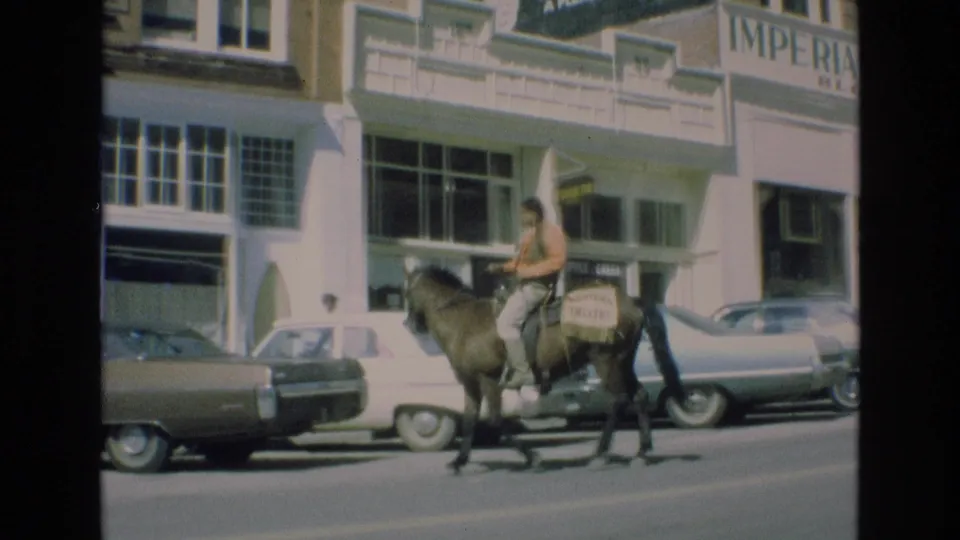 A sleeveless rider on horseback passes parked cars and commercial storefronts in downtown Colorado Springs, with hotel-block signage partly visible above the sidewalk.