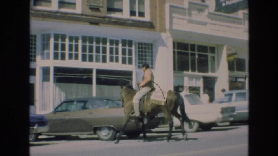 The mounted rider continues along the downtown Colorado Springs street in front of large storefront windows, with 1970s-era cars parked at the curb.