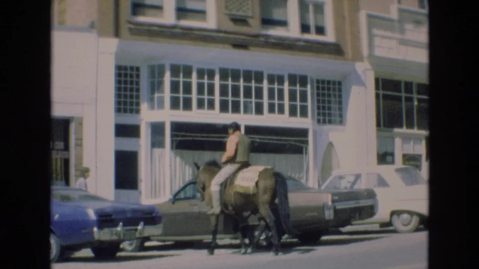 A man on horseback rides past parked cars and a storefront facade in downtown Colorado Springs, with the street scene framed from across the road.