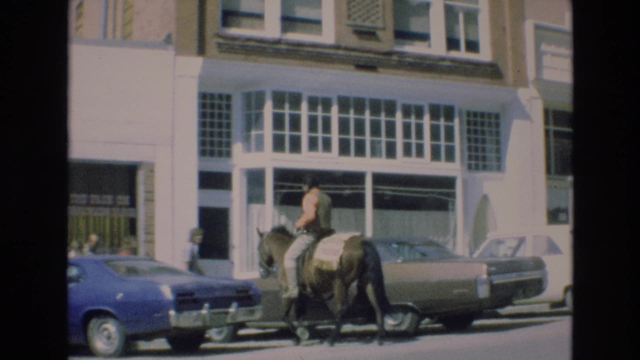 In the supplied 1973 Colorado Springs home-movie context, a rider on a dark horse moves along a city street past a blue car, a brown sedan, and large storefront windows.