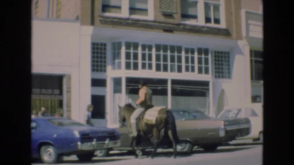 In the supplied 1973 Colorado Springs home-movie context, a rider on a dark horse moves along a city street past a blue car, a brown sedan, and large storefront windows.