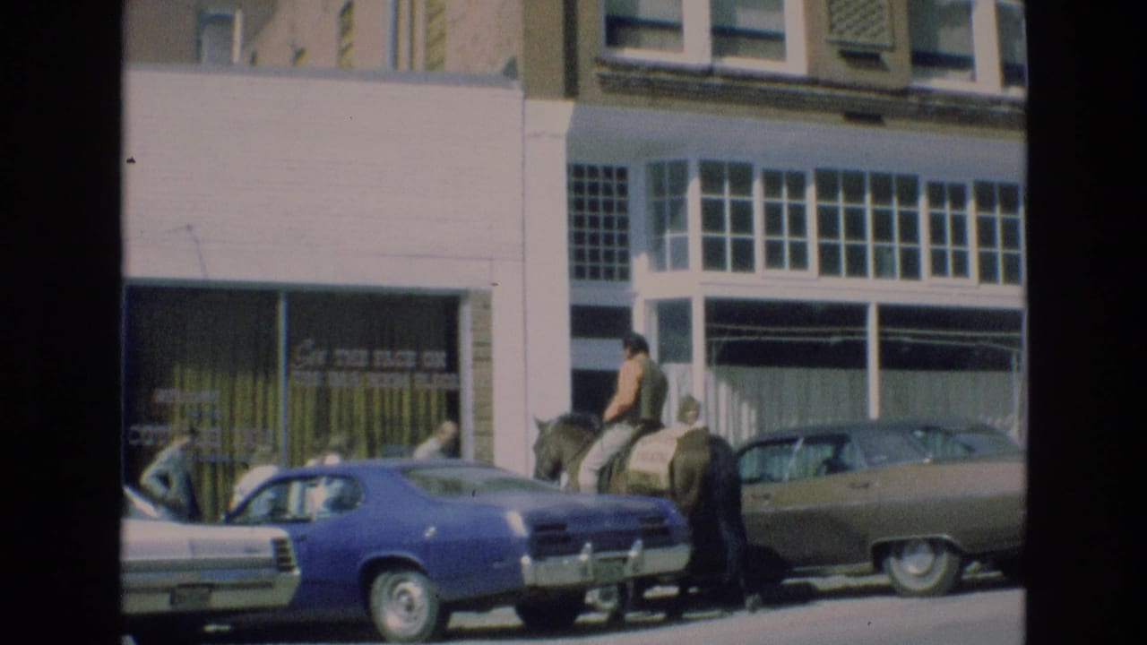 The rider continues along the street beside parked 1970s cars, with pedestrians and a storefront sign visible in the background.