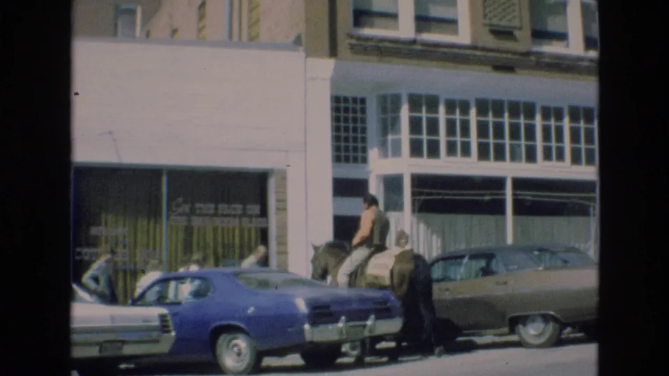The rider continues along the street beside parked 1970s cars, with pedestrians and a storefront sign visible in the background.
