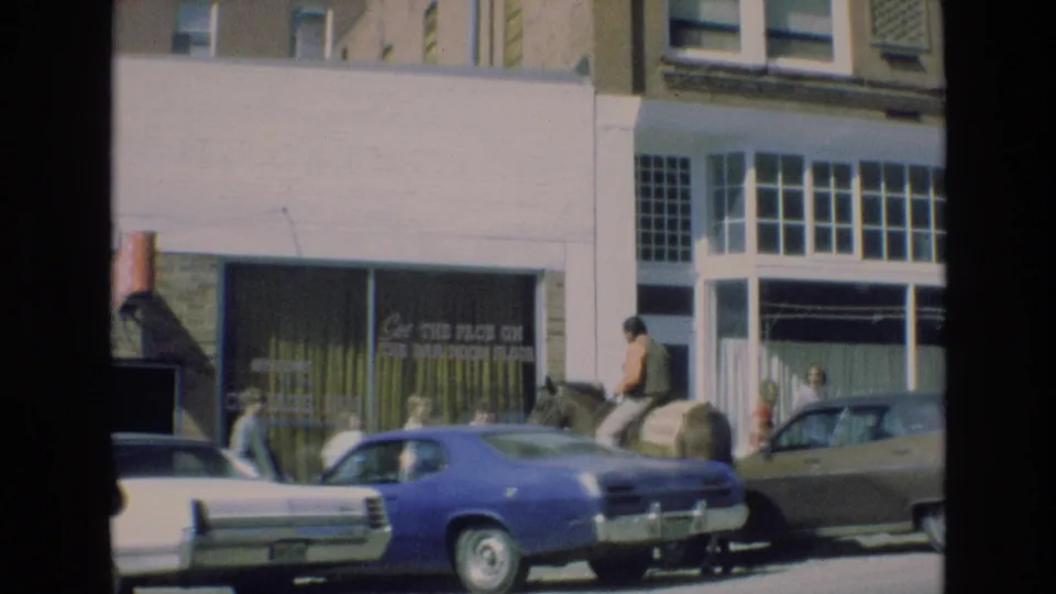 In a Colorado Springs street view from the supplied 1973 clip, the rider and horse pass a blue parked car while pedestrians stand near a storefront.