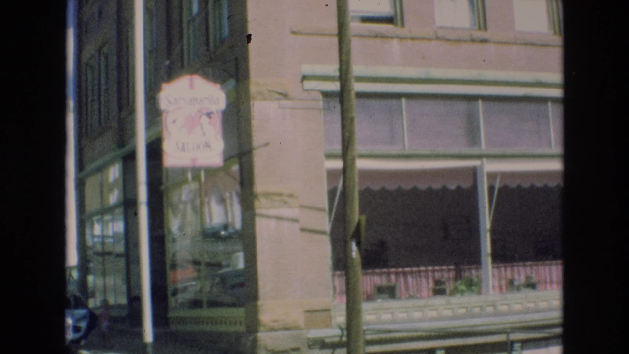 1973 Colorado Springs street view showing a pink Saraparilla Saloon sign mounted on a brick building with storefront glass, awning, and a utility pole in the foreground.
