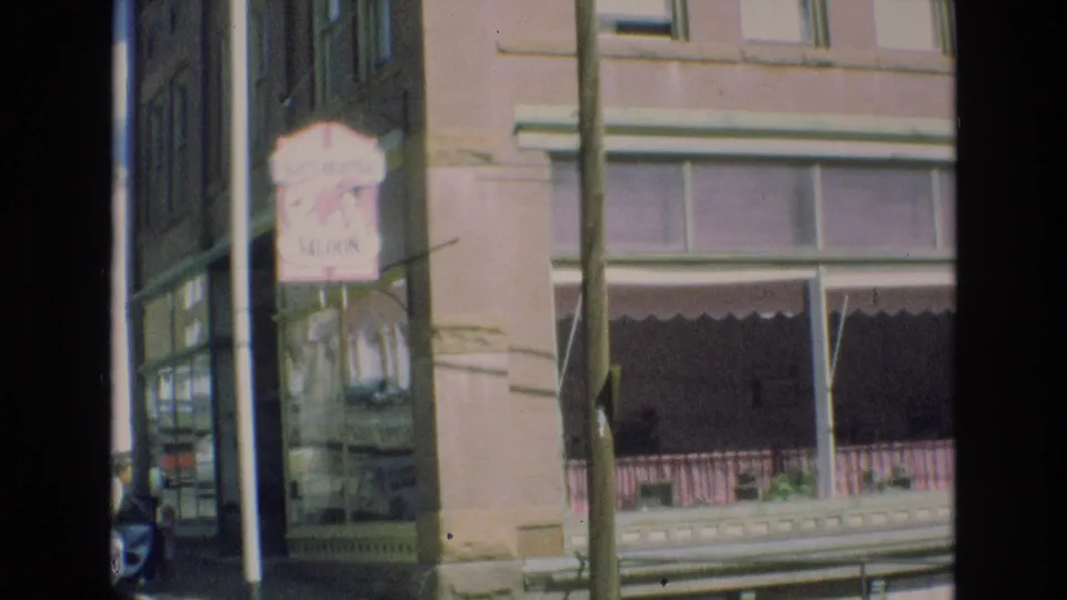 1973 Colorado Springs home-movie frame with a pink Saraparilla Saloon sign, brick commercial facade, large windows, and a foreground pole.