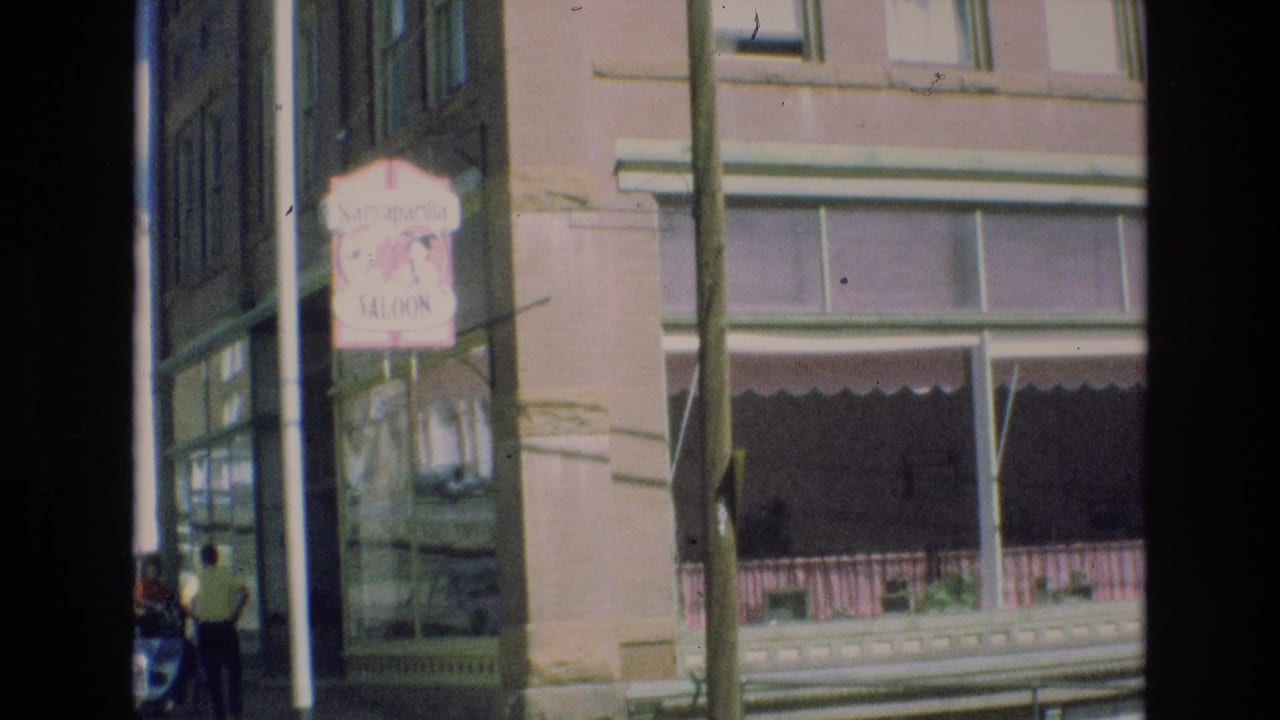 1973 Colorado Springs street view showing a Sarsaparilla Saloon sign on a corner building, a shaded storefront awning, sidewalk detail, and pedestrians partly visible at the frame edge.