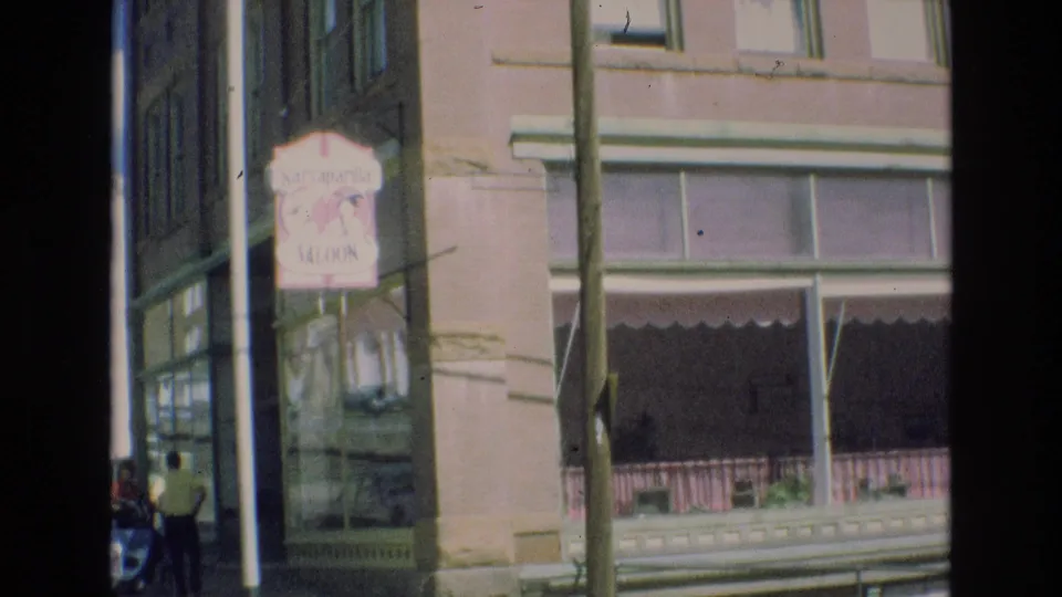 1973 Colorado Springs street view showing a Sarsaparilla Saloon sign on a corner building, a shaded storefront awning, sidewalk detail, and pedestrians partly visible at the frame edge.