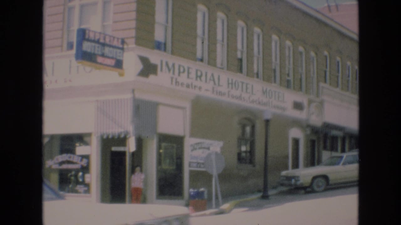 1973 Colorado Springs home-movie frame showing the Imperial Hotel-Motel sign, storefront entrance, a woman at the sidewalk, and classic cars parked beside the building.