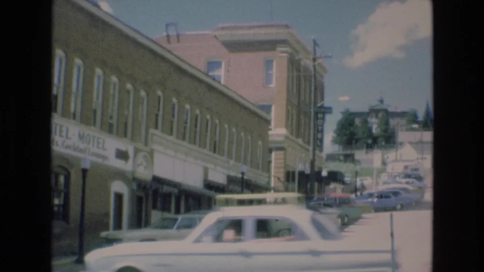 1973 Colorado Springs frame with a moving car crossing the foreground, the Imperial Hotel-Motel facade behind it, and a line of parked cars continuing along the block.