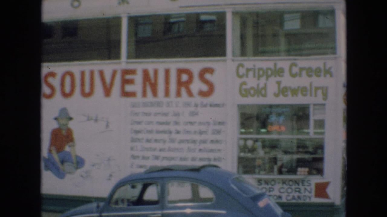 1973 Colorado Springs storefront view showing red SOUVENIRS lettering, Cripple Creek Gold Jewelry signage, display windows, and a blue compact car at the curb.
