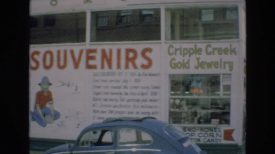 1973 Colorado Springs storefront view showing red SOUVENIRS lettering, Cripple Creek Gold Jewelry signage, display windows, and a blue compact car at the curb.