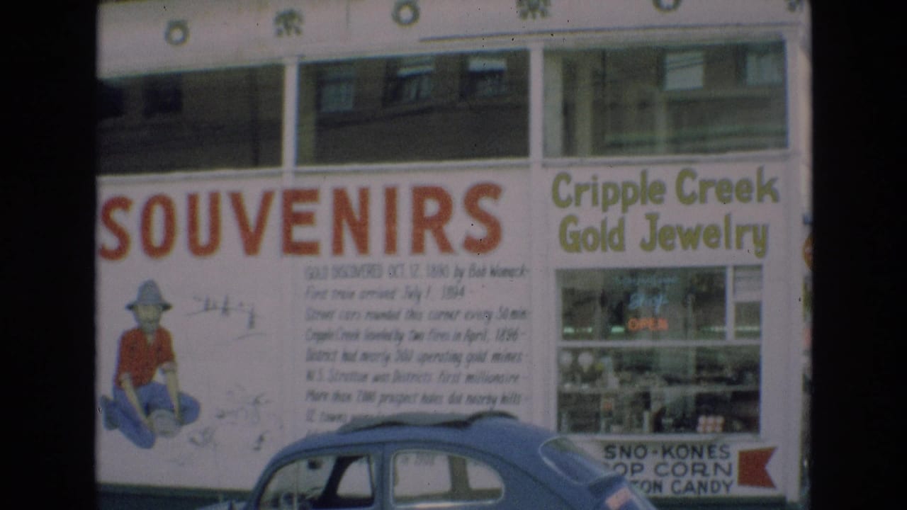 1973 Colorado Springs frame showing a souvenir sign with a prospector illustration, adjacent Cripple Creek Gold Jewelry storefront, lit OPEN sign, and blue car parked along the street.