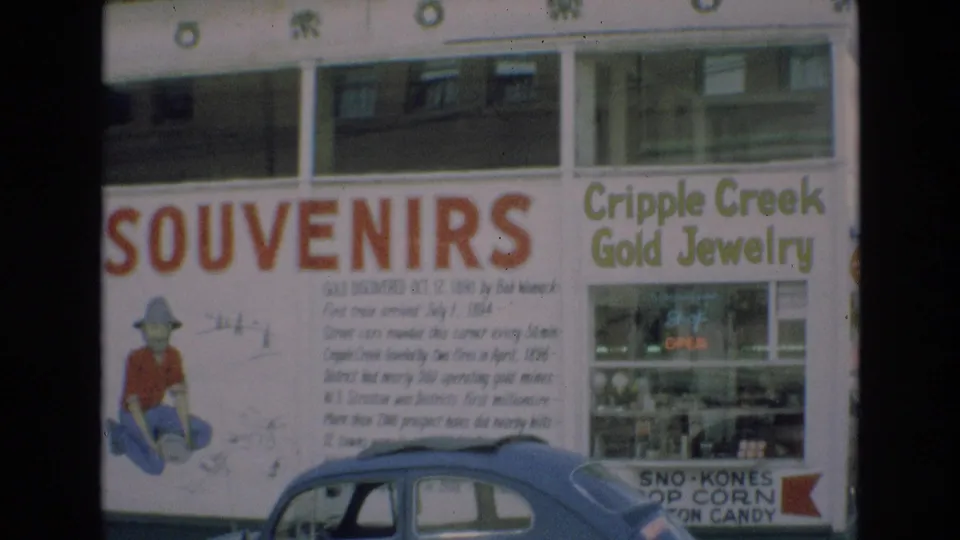 1973 Colorado Springs frame showing a souvenir sign with a prospector illustration, adjacent Cripple Creek Gold Jewelry storefront, lit OPEN sign, and blue car parked along the street.