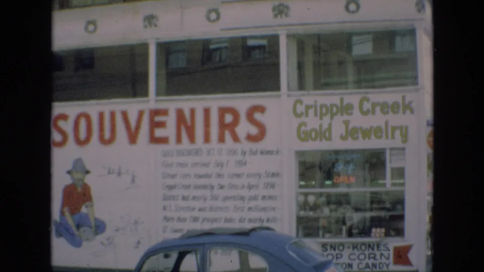1973 Colorado Springs home-movie frame with prominent SOUVENIRS lettering, prospector graphic, Cripple Creek Gold Jewelry sign, and a blue compact car along the bottom edge.