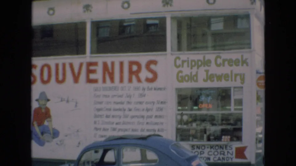 1973 Colorado Springs storefront frame showing Western-themed souvenir signage, Cripple Creek Gold Jewelry lettering, window displays, a small OPEN sign, and a blue car in front.