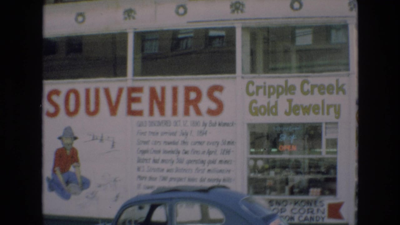 1973 Colorado home-movie frame showing a souvenir shop facade with Cripple Creek Gold Jewelry lettering, historical gold-discovery text, and a parked car partly blocking the lower storefront.