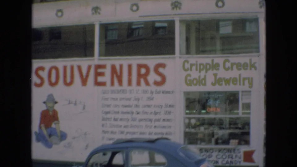1973 Colorado home-movie frame showing a souvenir shop facade with Cripple Creek Gold Jewelry lettering, historical gold-discovery text, and a parked car partly blocking the lower storefront.