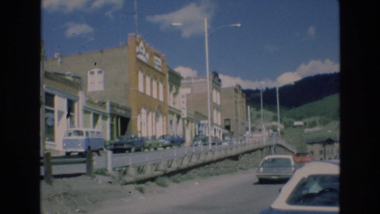 Quiet main street with parked cars, storefronts, streetlamps, and mountains beyond.