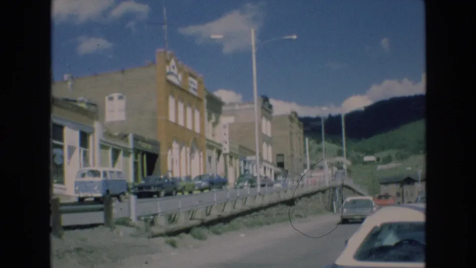 1973 Colorado home-movie frame showing a roadside view past older storefronts, a blue Volkswagen van, several parked cars, a sloped street edge, and mountains under a bright sky.