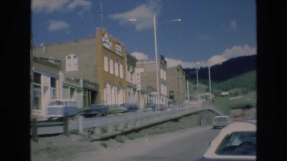1973 Colorado home-movie frame capturing a quiet commercial street with older buildings, parked vehicles, upright streetlights, a roadside barrier, and green hills beyond the town.