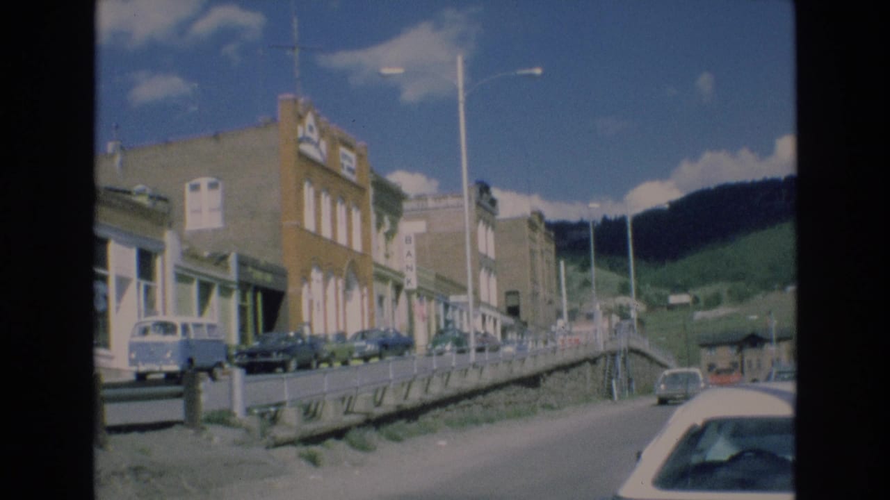 1973 Colorado Springs street view showing commercial buildings, parked cars, a blue-and-white van, street lamps, and green hills beyond the road.