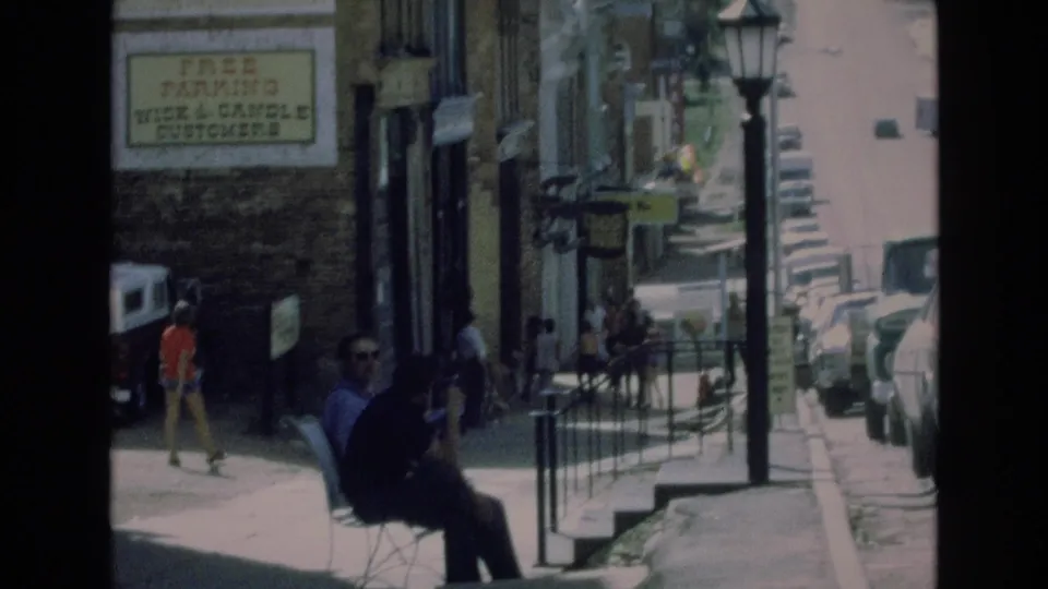 1973 Colorado Springs sidewalk view showing pedestrians, a seated person near a bench, storefront windows, railings, parked cars, and a free-parking sign for Wick & Candle customers.