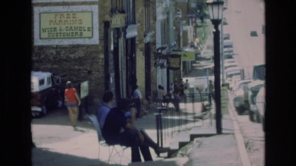 1973 Colorado Springs sidewalk scene with pedestrians, a seated figure, storefronts, a visible Wick & Candle customer parking sign, railings, parked cars, and sunlit pavement.