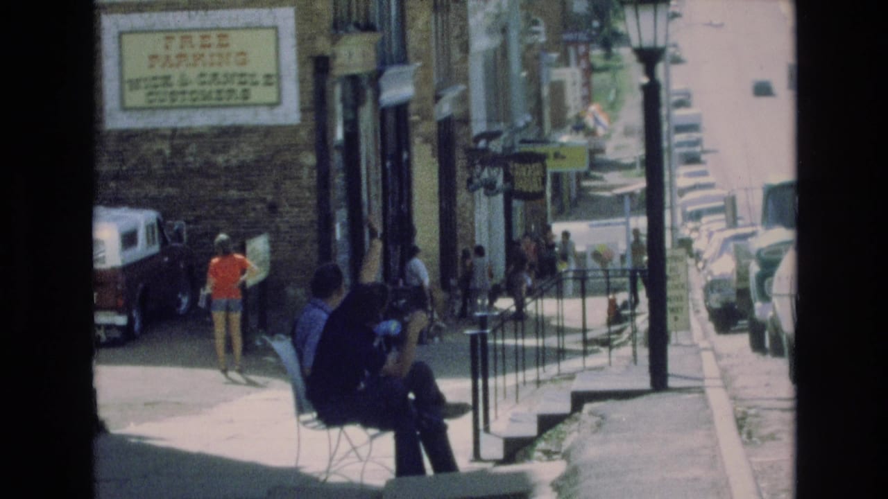 1973 Colorado Springs sidewalk view with seated figures in the foreground, a person in an orange top walking past a brick storefront, parked cars, railings, and a free-parking sign.