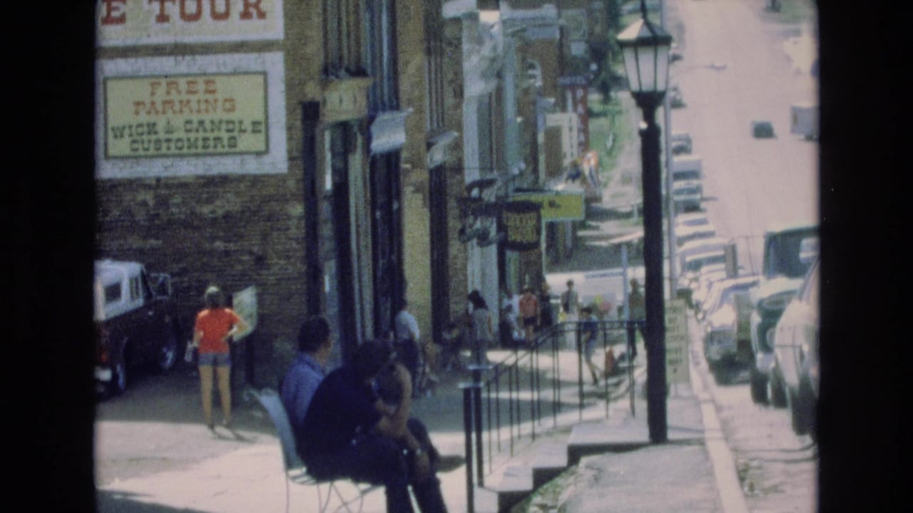 1973 Colorado Springs street scene showing pedestrians along a storefront sidewalk, a seated person in dark clothing, parked cars, railings, a streetlamp, and a Wick and Candle customer parking sign.
