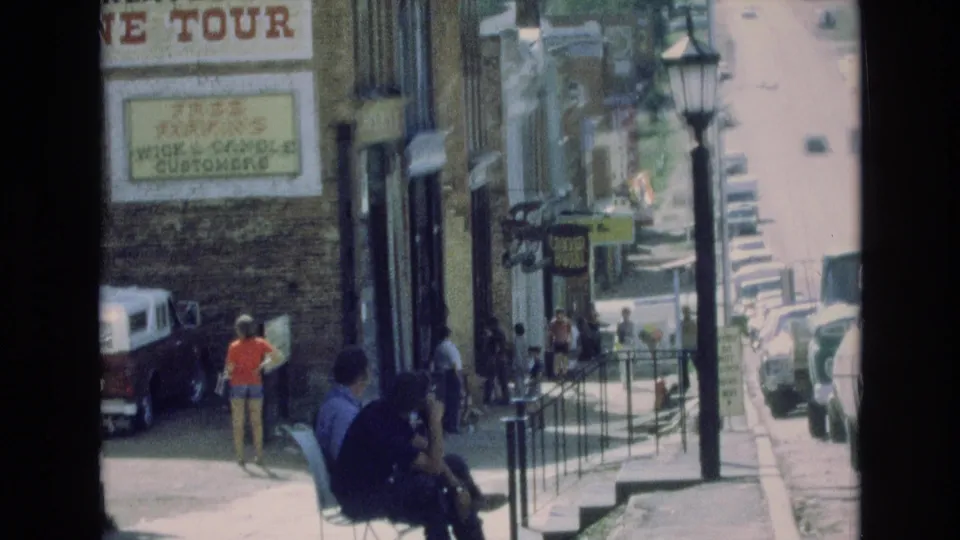 1973 Colorado Springs home-movie frame with seated sidewalk figures, a pedestrian in orange near a parked vehicle, storefront signage, railings, steps, and a sunlit street receding in the background.