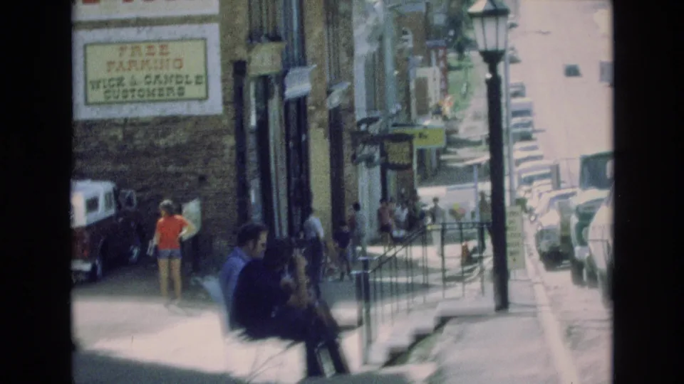 1973 Colorado Springs sidewalk frame with seated figures in the foreground, pedestrians near shop entrances, parked cars along the curb, a streetlamp, railings, and a free-parking sign on a brick building.