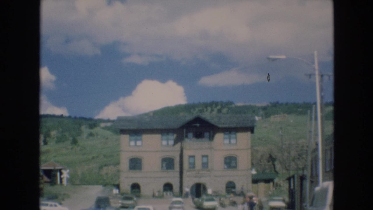 1973 Colorado Springs home-movie frame showing a two-story building with arched windows, parked cars, green hills, utility poles, and a blue sky with clouds.