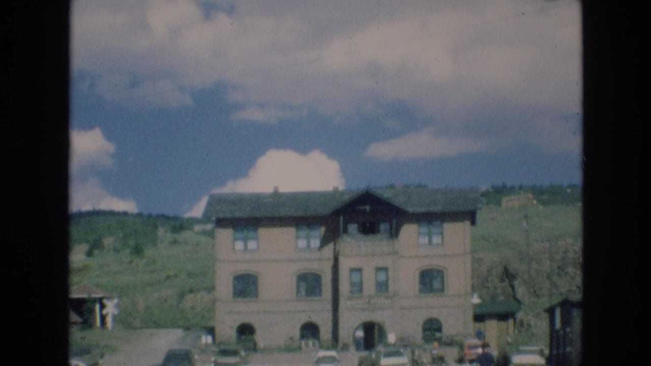 Building facade with parked cars, distant pedestrians, hillside, and cloudy sky.