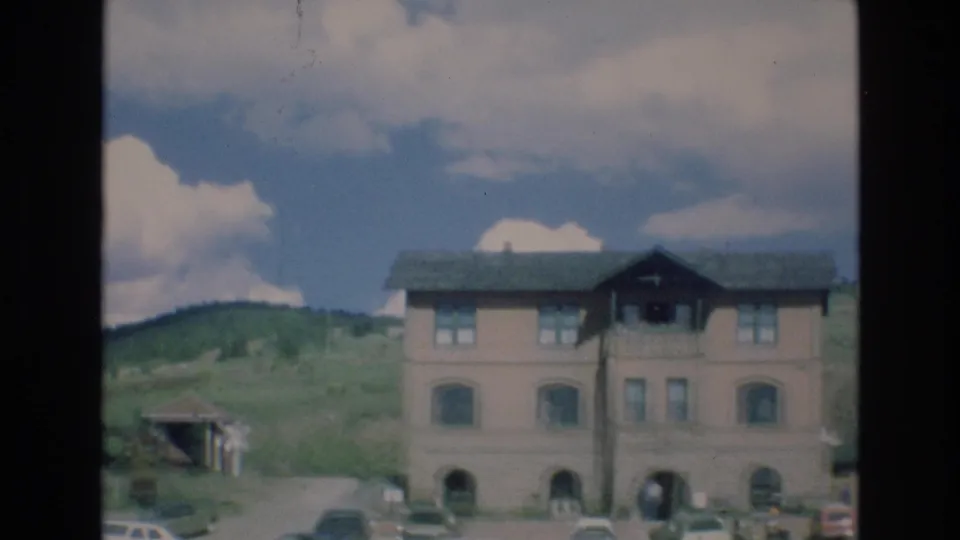 1973 Colorado Springs home-movie frame with the building partly centered, parked cars along the road, a small roadside structure, green hills, and large clouds.
