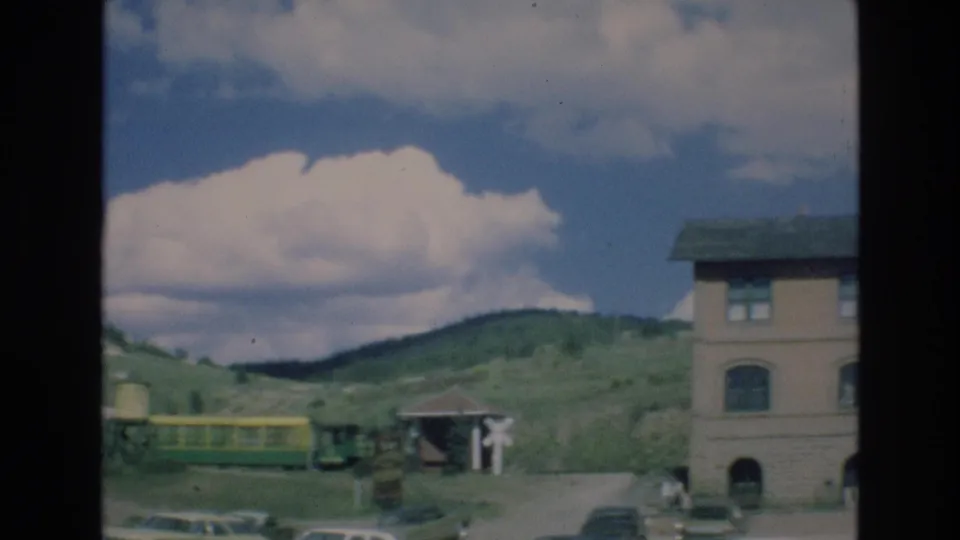 1973 Colorado Springs frame showing a railroad crossing area with a yellow and green train car, parked cars, a small structure, green hills, clouds, and the edge of a building.