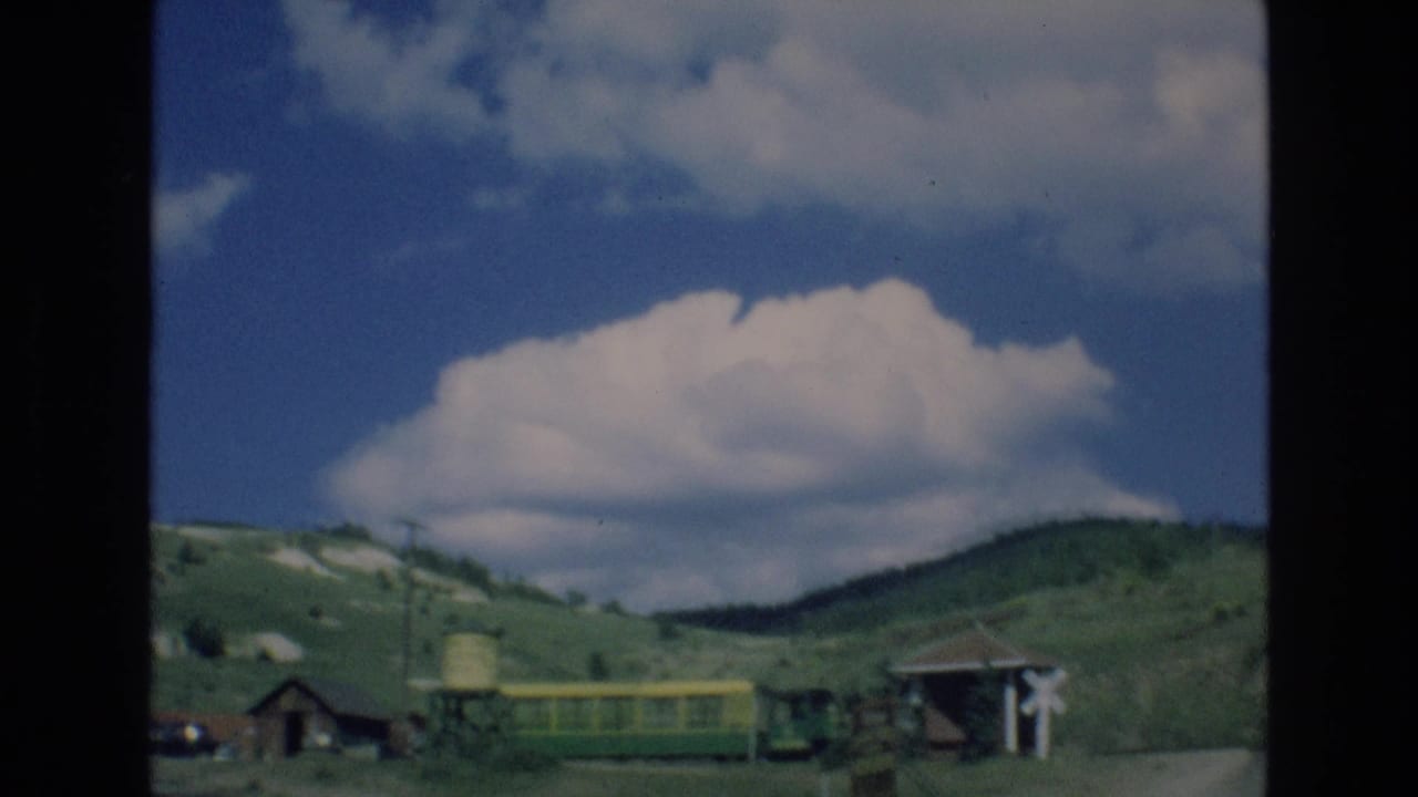 1973 Colorado Springs home-movie frame showing a mountain village scene with rolling green hills, rustic structures, a water tower, and a green-and-yellow tourist train or tram beneath large white clouds.