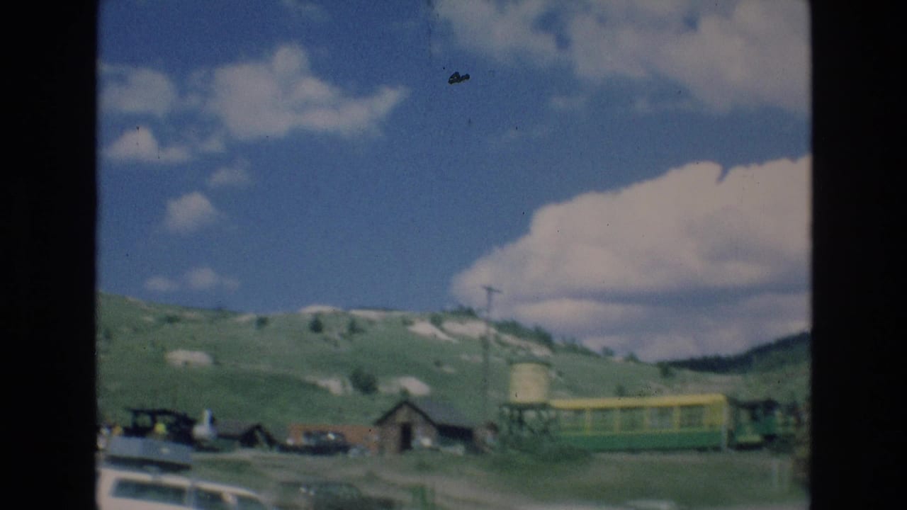 Tourist train, rustic buildings, road edge, water tower, and open hillside beneath clouds.