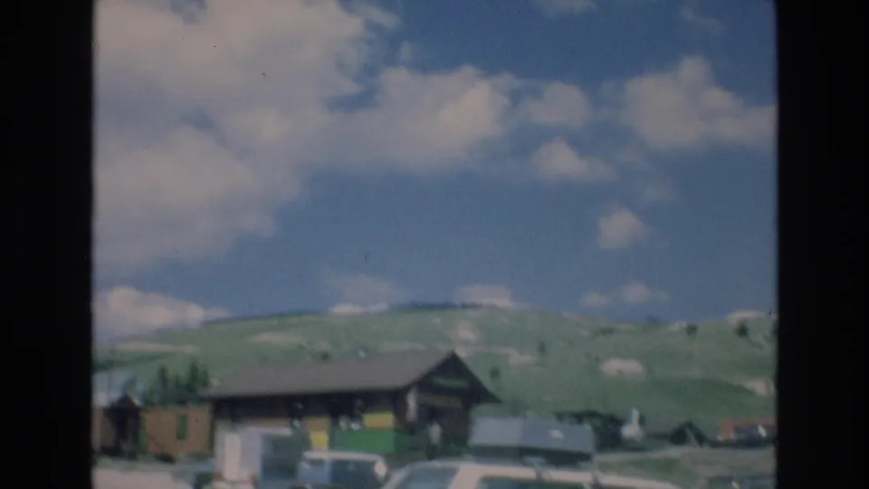 1973 Colorado Springs home-movie view of a rustic wooden building and parked period cars along a roadside village, with green hills and scattered clouds in the background.