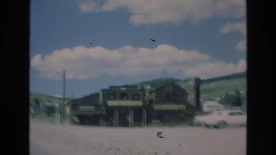 1973 Colorado Springs frame showing a dirt road beside rustic wooden buildings, a railroad crossing sign, a parked 1970s car, and rolling hills under a bright clouded sky.
