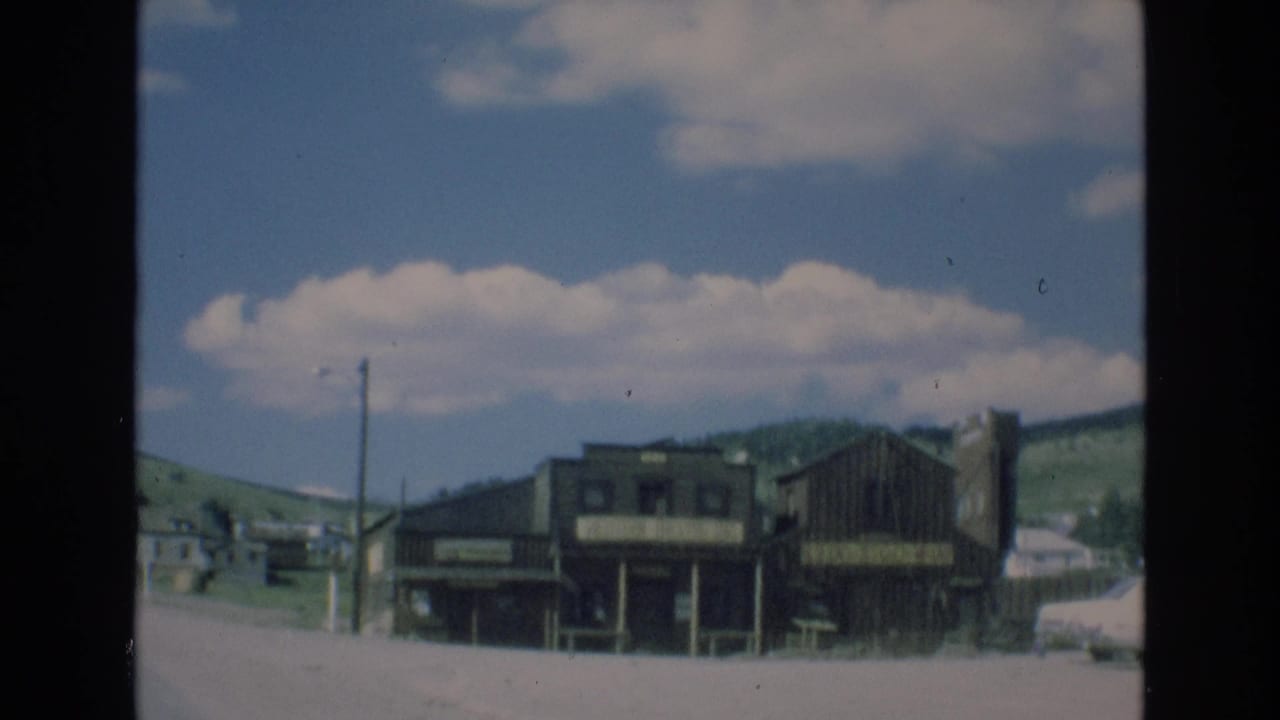 1973 Colorado-area 8mm frame showing a quiet street with weathered wooden storefront-style buildings, utility poles, low hills, and scattered clouds.
