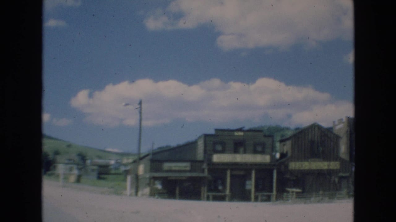 Roadside wooden buildings and utility poles beneath a bright cloudy sky.