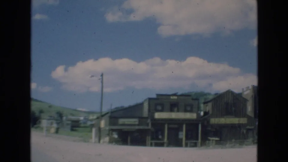 1973 Colorado-area 8mm frame with roadside wooden buildings, a utility pole, open street space, distant hills, and a bright sky with scattered clouds.