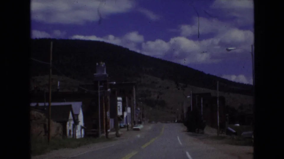 1973 Colorado-area 8mm frame looking down a two-lane road through a quiet small town, with storefronts, utility poles, a hillside, and clouded sky.